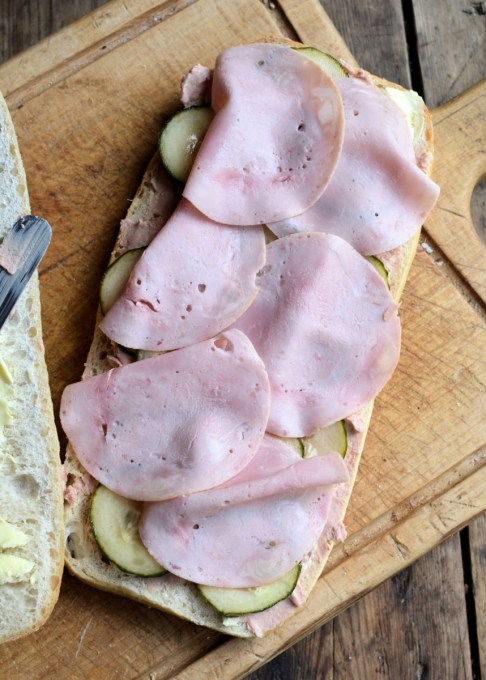 Method: 1. Cut the loaf of bread in half as seen in the photos. 2. Spread both halves of the bread with the butter and then layer up the ingredients on one half of the bread loaf in the order listed – see the photos. 3. Place the lid on top of the filling and then wrap in greaseproof paper and tie with some string. 4. Place some heavy books or weights on top and leave for at least 12 to 24 hours, in a cool place. 5. Serve in slices.