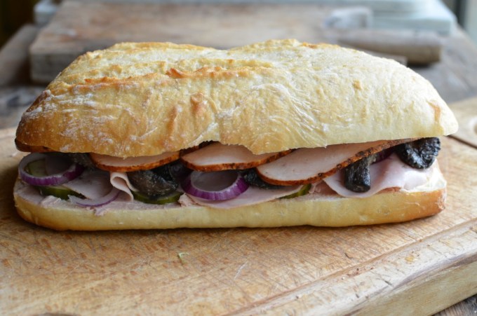 Method: 1. Cut the loaf of bread in half as seen in the photos. 2. Spread both halves of the bread with the butter and then layer up the ingredients on one half of the bread loaf in the order listed – see the photos. 3. Place the lid on top of the filling and then wrap in greaseproof paper and tie with some string. 4. Place some heavy books or weights on top and leave for at least 12 to 24 hours, in a cool place. 5. Serve in slices.